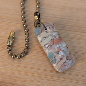 May include: A rectangular keychain with a mosaic design in shades of red, beige, and blue-green. It is attached to a brass-colored ball chain and a clasp. The keychain is likely handmade and unique.