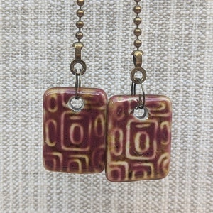 May include: Two square, burgundy-colored ceramic pendants with a repeating geometric pattern in a lighter beige hue. Each pendant is attached to a bronze-colored ball chain. The pendants are hanging against a neutral, textured background.