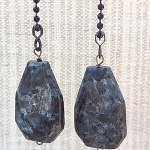 May include: A pair of earrings featuring dark gray, faceted, coffin-shaped labradorite stones suspended from black metal ball chains. Each stone displays iridescent blue and silver inclusions within the dark, polished mineral surface.