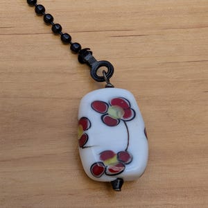 May include: A white rectangular glass bead pendant featuring a floral pattern with dark red petals and yellow centers. The bead is attached to a black metal ball chain by a dark metal loop, creating a functional pull for a ceiling fan or light.