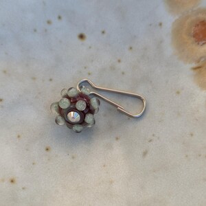 May include: A stitch marker featuring a spherical dark purple glass bead adorned with small, raised pale green dots and a central silver-toned metal cone accent, attached to a silver-toned metal bulb-shaped safety pin clasp for knitting projects.