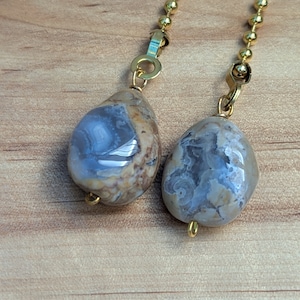 May include: Two agate stone ceiling fan pulls with gold-colored hardware. The stones are oval-shaped, with a mix of beige, brown, and blue-gray colors. The pulls hang from a gold ball chain.