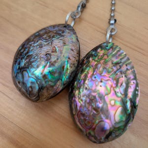 May include: Two teardrop-shaped abalone shell pendants with iridescent surfaces displaying swirls of green, blue, purple, and pink. Each pendant is attached to a silver-toned metal ball chain by a small circular jump ring, creating a decorative pull.