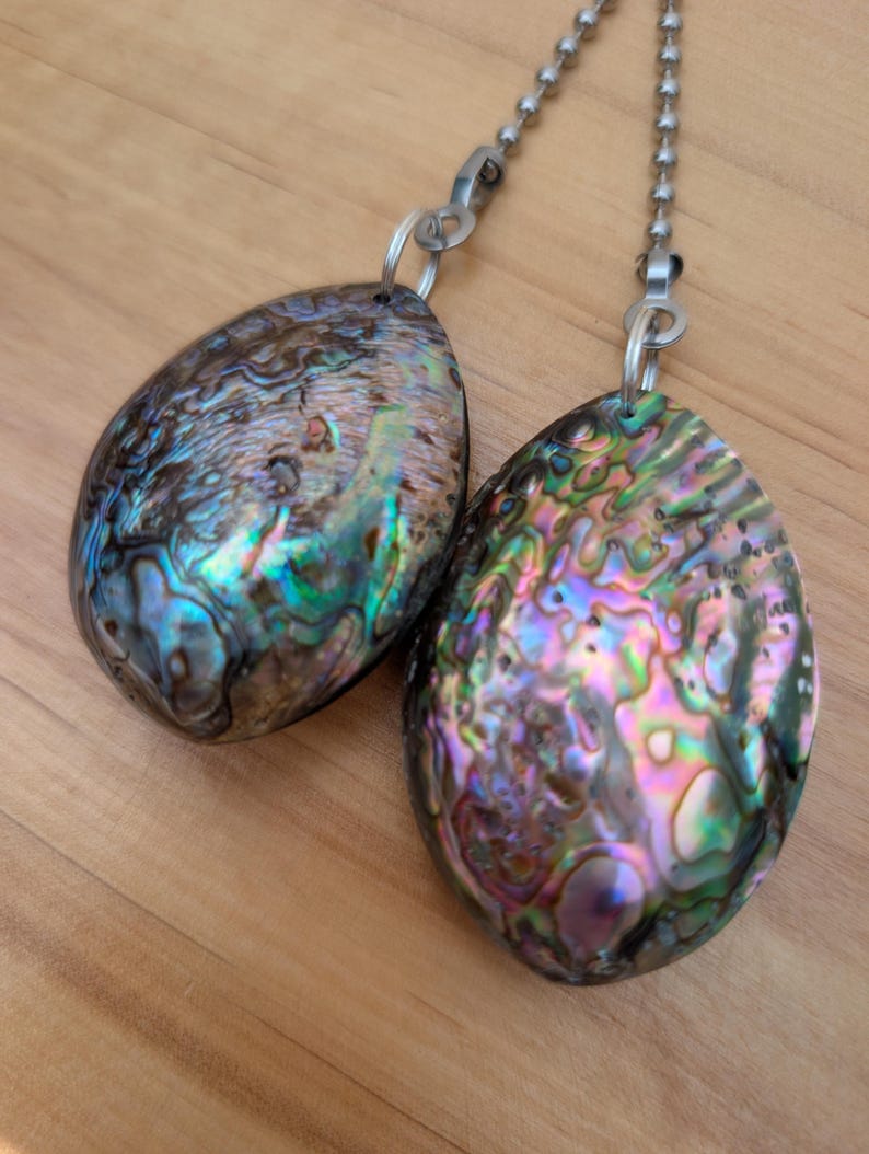 May include: Two teardrop-shaped abalone shell pendants with iridescent surfaces displaying swirls of green, blue, purple, and pink. Each pendant is attached to a silver-toned metal ball chain by a small circular jump ring, creating a decorative pull.