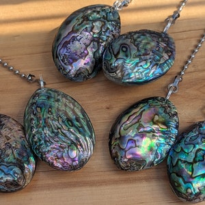 May include: Five oval-shaped abalone shell pendants, each featuring iridescent swirls of green, blue, purple, and pink, are attached to silver-toned metal ball chains. The shells display natural, unique patterns with a polished, glossy finish.