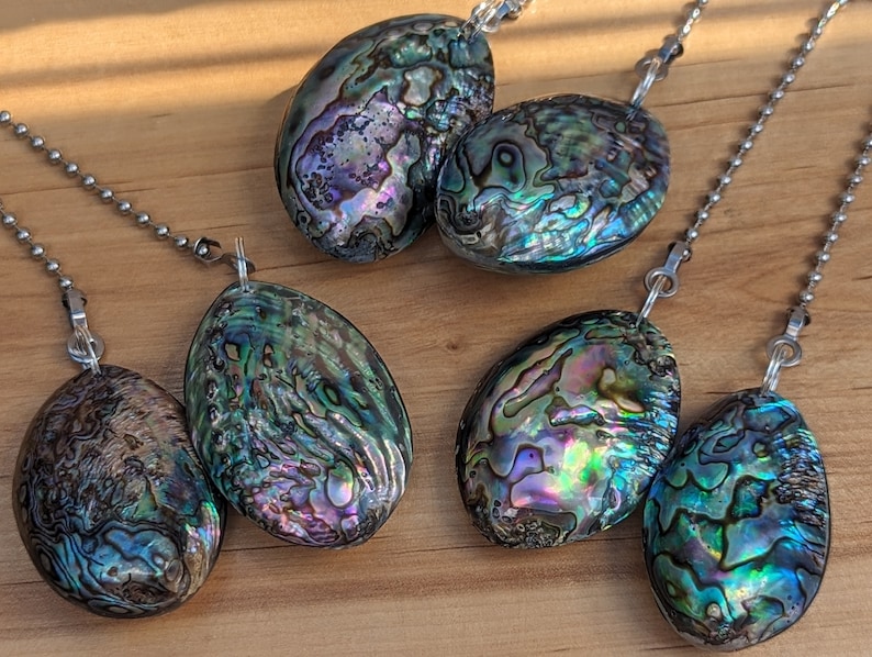 May include: Five oval-shaped abalone shell pendants, each featuring iridescent swirls of green, blue, purple, and pink, are attached to silver-toned metal ball chains. The shells display natural, unique patterns with a polished, glossy finish.