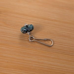 May include: A decorative charm featuring a round, swirled glass bead in shades of teal, black, and white, attached to a silver-toned metal lobster claw clasp. The bead has a smooth, glossy finish and is connected to the clasp by a small metal ring.