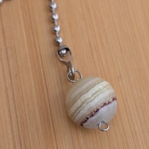 May include: A decorative ceiling fan pull chain with a striped, spherical, off-white and tan bead. The bead has a thin band of maroon near the bottom. The chain is made of small, silver-colored ball links.