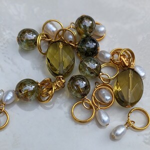 May include: Close-up of jewelry components featuring olive green faceted beads, gold-colored rings, and white pearl accents. The beads have a speckled appearance. The components are arranged on a light-colored surface.