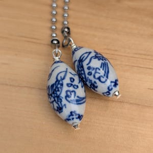May include: Two white and blue porcelain light pull chain ornaments. Each ornament is an elongated oval shape with blue floral designs. The ornaments are attached to a silver ball chain.