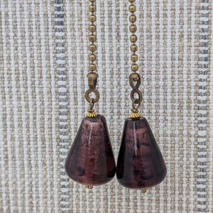 May include: Two ceiling fan pulls featuring teardrop-shaped glass beads in a deep plum color with metallic flecks. Each bead is attached to a brass-toned metal ball chain by a small jump ring and a decorative gold-toned cap at the top and bottom.