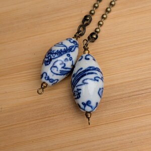 May include: Two oval-shaped ceramic beads featuring a white base with intricate blue floral patterns. Each bead is attached to a bronze-toned metal ball chain connector with a small loop at the bottom, creating a decorative pull for a light fixture.