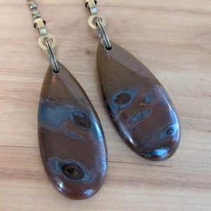 May include: Two teardrop-shaped brown stone pendants with dark blue and black swirling patterns, each attached to a metallic ball chain connector by a small silver-toned jump ring. The stones have a smooth, polished, and glossy finish.