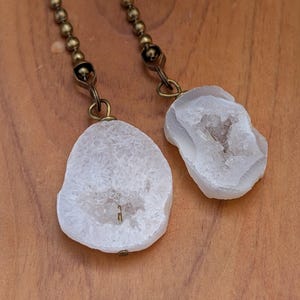 May include: Two white geode slices with crystalline centers attached to antique brass ball chains. Each irregular stone features a rough, textured surface with a sparkling, hollow core, suspended by a small metal loop connected to the chain.