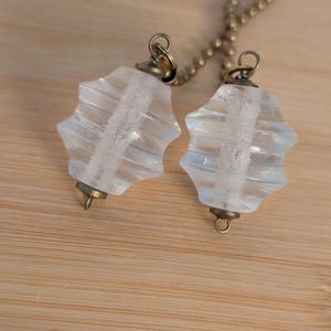 May include: Two clear, star-shaped glass pendants with brass-colored metal accents and a delicate chain. The pendants have a ridged design and are suspended against a light brown wooden surface.