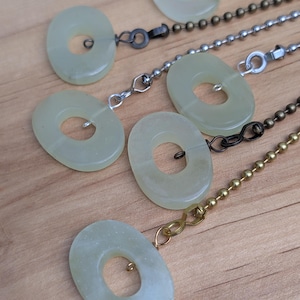 May include: Five pale green, oval-shaped stone pulls attached to various metal ball chains. Each pull features a central circular cutout and is connected to a chain using a small metal jump ring. The chains vary in color, including silver, brass, and dark bronze tones.