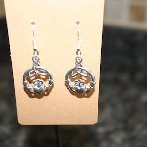 May include: A pair of silver-toned Claddagh earrings. Each earring features a heart, crown, and hands design within a circular frame, suspended from a simple hook. The earrings are displayed on a tan card.