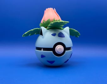 May include: A blue and white Poke Ball with a green and pink flower on top. The Poke Ball is a popular collectible toy from the Pokemon franchise.