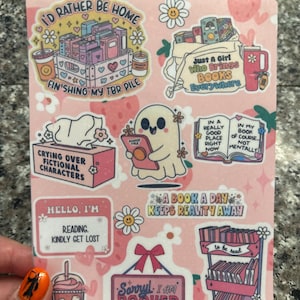 Peut inclure: Un ensemble de 10 autocollants roses et blancs avec des illustrations et du texte sur le thème du livre. Les autocollants présentent des phrases comme "Je préférerais être à la maison", "Juste une fille qui apporte des livres partout", "Pleurer sur des personnages fictifs", "Bonjour, je lis. Veuillez vous perdre", "Un livre par jour éloigne la réalité", "Désolé, je suis réservé. Veuillez me laisser tranquille", et "À lire".