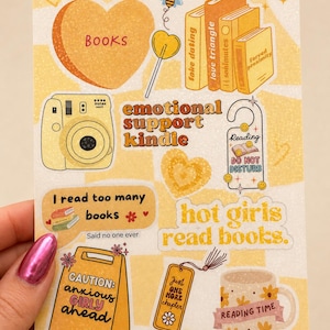 May include: A sticker sheet featuring yellow and orange illustrations including a heart labelled BOOKS, a stack of books, an Instax camera, a door hanger reading READING DO NOT DISTURB, and text including emotional support kindle and hot girls read books.