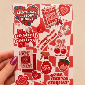 May include: A sticker sheet featuring various red and pink designs including a heart with text Emotional Support Kindle, a book labelled I Like My Books Extra Spicy, dice, cherries, a spray bottle labelled Book Spoiler Repellent, and a strawberry.