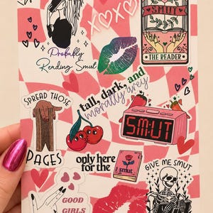 May include: A sticker sheet featuring a pink and white checkered background with various illustrations including a tarot card labelled The Reader, a digital clock displaying SM:UT, a skeleton reading a book, cherries, lips, and text phrases.