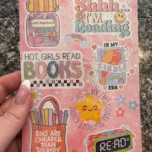 May include: A pink sheet of stickers with a variety of designs related to reading. The stickers feature phrases like "Shh... I'm Reading", "Hot Girls Read Books", "Books Are Cheaper Than Therapy", and "This Is My Emotional Kindle Support".
