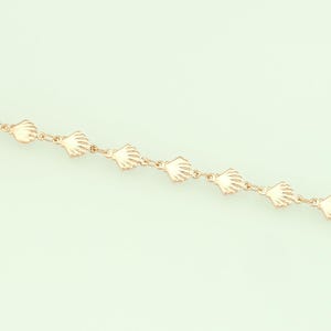 Puede incluir: Una delicada pulsera de cadena dorada con un patrón repetido de pequeños dijes de conchas marinas. La pulsera se muestra en diagonal sobre un fondo verde claro y suave, mostrando su intrincado diseño y tema costero.