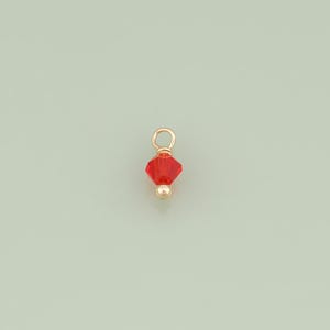 May include: A small jewelry charm featuring a faceted, bicone-shaped red crystal bead suspended from a gold-toned metal jump ring, with a smooth, round gold-toned metal bead positioned at the base of the crystal to complete the pendant design.