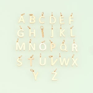 May include: A collection of gold-colored alphabet letter charms, each with a small loop at the top for attaching to a necklace or bracelet. The letters are arranged in rows and columns against a light green background.
