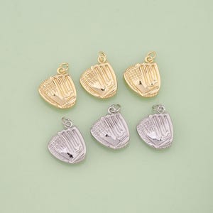May include: Six baseball glove charms in gold and silver tones. The charms are detailed with stitching and a loop for attaching to jewelry. The gloves are arranged on a light green background.