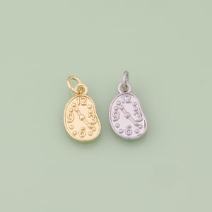 May include: Two small, gold and silver-colored clock charms. Each charm has a rounded, melting clock face design with the number 12 at the top. The clock hands point to the time. The charms are on a light green background.