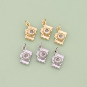 Op de afbeelding: Zes kleine camera bedels in goud- en zilverkleuren op een lichtgroene achtergrond. Elke bedel heeft een rechthoekig lichaam met een cirkelvormige lens en een kleine lus om te bevestigen. De bedels zijn gedetailleerd en glanzend.