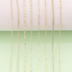 May include: A selection of gold-coloured necklaces featuring diverse chain styles. The chains include delicate box, rope, and beaded designs, presented against a soft white and green backdrop. These necklaces are likely for jewellery or fashion.