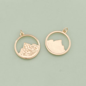May include: Two gold-toned metal charms in the shape of a circle with a mountain range design inside. One charm has a solid mountain range design, while the other has an outline of the mountain range.