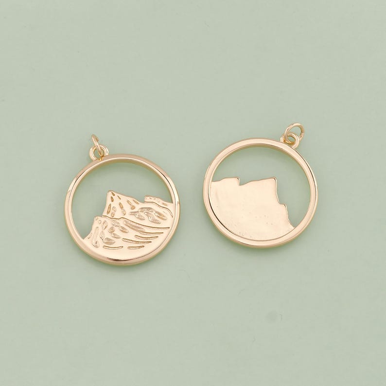 May include: Two gold-toned metal charms in the shape of a circle with a mountain range design inside. One charm has a solid mountain range design, while the other has an outline of the mountain range.