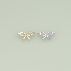 May include: Two bow-shaped rings, one gold-colored and one silver-colored, are displayed against a light green background. The rings have a delicate, minimalist design, with the bow detail as the focal point. These rings are suitable for everyday wear.