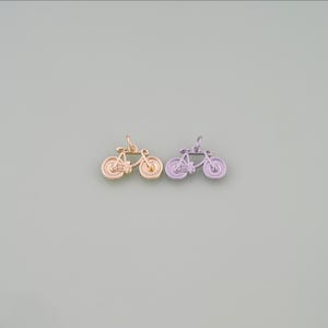 May include: Two small bicycle charms, one in a metallic gold finish and one in a metallic silver finish. Each charm features a detailed frame, handlebars, and two wheels with a textured spoke design, complete with a small jump ring at the top.