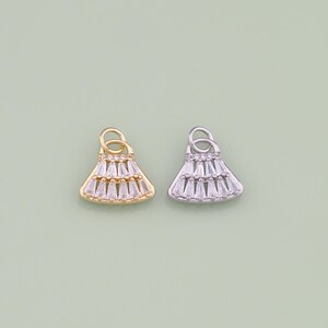 May include: Two small, decorative charms. One is gold-toned, the other silver-toned. Both charms are shaped like a tiered skirt, with clear, rectangular stones. Each charm has a small loop at the top for attaching to jewelry. The background is a light green color.