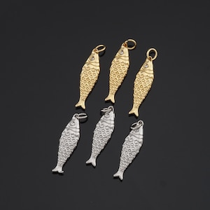 May include: Six fish-shaped pendants in gold and silver tones. The pendants have a textured scale design and a small loop at the top for attaching to a chain or other jewelry. The gold pendants have a small clear stone at the top.