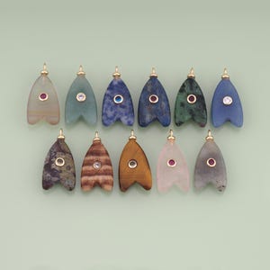 May include: Eleven teardrop-shaped stone pendants arranged in two rows. Each pendant features a gold-tone metal bail at the top and a small, circular, bezel-set gemstone at the centre. The stones vary in colour, including translucent, blue, green, brown, and pink hues.