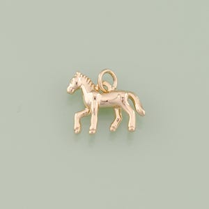 May include: A gold-colored horse charm with a loop for attaching to a necklace or bracelet. The horse is depicted in a walking pose, with detailed legs, mane, and tail. The charm is likely made of metal and has a polished finish.
