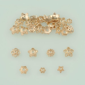 May include: A collection of gold-colored metal flower-shaped bead caps in various sizes and designs. The bead caps have detailed floral patterns and are ideal for jewelry making. The background is a light teal color.