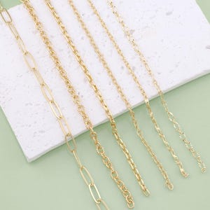 May include: Six gold chains of different styles and lengths laid out on a white surface. The chains are all made of a shiny gold metal.
