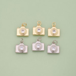 Op de afbeelding: Zes kleine camera bedels in goud en zilver. Elke bedel heeft een centrale heldere steen en een lus om aan sieraden te bevestigen. De camera's zijn gerangschikt in twee rijen van drie.