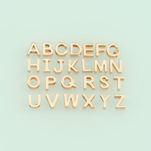 May include: Gold-colored metal alphabet letter charms, A through Z, arranged in three rows on a light green background.