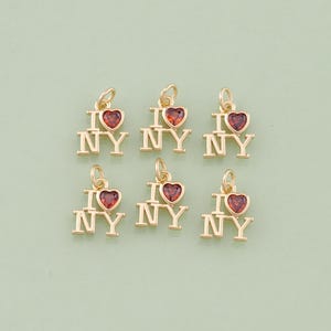 May include: Six gold-colored charms with the phrase "I NY" and a red heart. The charms are small and detailed, with a loop at the top for attaching to a necklace or bracelet. The background is a solid light green.