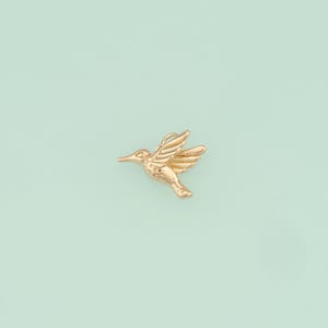 Puede incluir: Un colgante de colibrí de color dorado. El colibrí está en vuelo con las alas extendidas.