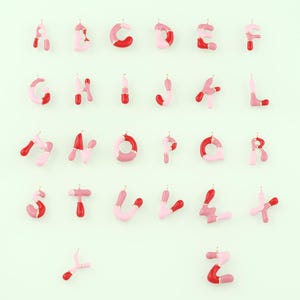 May include: Alphabet letter charms in a variety of pink and red hues, with gold-colored metal accents. Each letter is a unique design, with a glossy finish. The letters are arranged on a light green background.