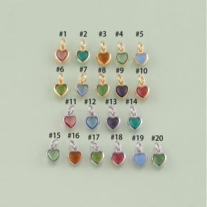 Pode incluir: Vinte pingentes em forma de coração em várias cores, incluindo vermelho, verde, azul e roxo. Cada pingente é numerado e tem um pequeno laço para prender a uma corrente ou pulseira. Os pingentes são exibidos em um fundo verde claro.